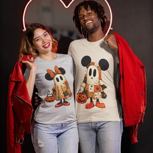 以下が含まれることがあります： ハロウィーンをテーマにしたお揃いのTシャツを着た2人。シャツには、ミッキーマウスの耳を持つ漫画の幽霊のキャラクターが描かれており、カボチャを持っています。幽霊はパッチワークのような衣装を着ています。背景は黒で、ネオンのハートがあります。