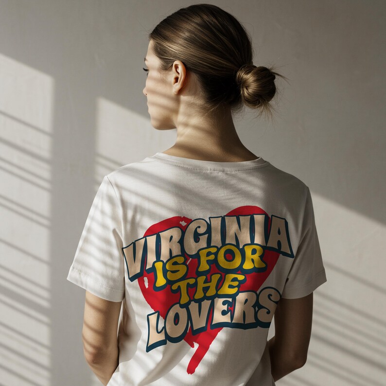 May include: Cream-colored t-shirt with the text "VIRGINIA IS FOR THE LOVERS" in a retro design. The text is set against a red heart shape with yellow and blue accents. The shirt is short-sleeved.