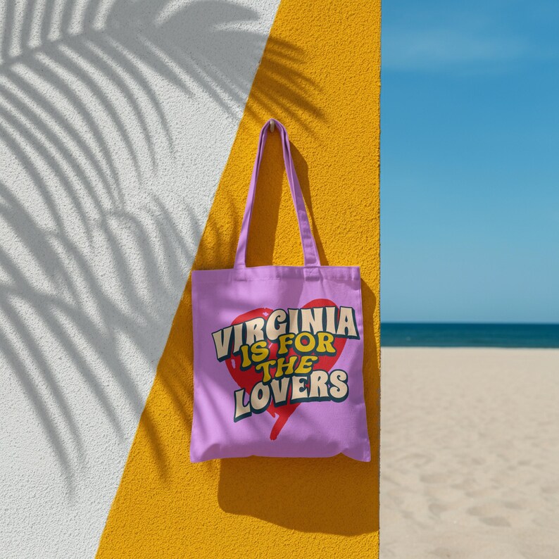 May include: A light purple tote bag with the text "VIRGINIA IS FOR THE LOVERS" in a heart-shaped design. The bag is hanging against a yellow wall with a white shadow and a blue sky and beach in the background.