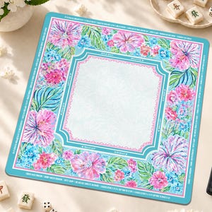 Op de afbeelding: Een vierkante mahjongmat met een lichtblauw middenstuk met een subtiel bladpatroon, omringd door een rand van roze hibiscusbloemen, blauwe hortensia's en groene bladeren. De buitenrand bevat witte tekstinstructies op een groenblauwe achtergrond.