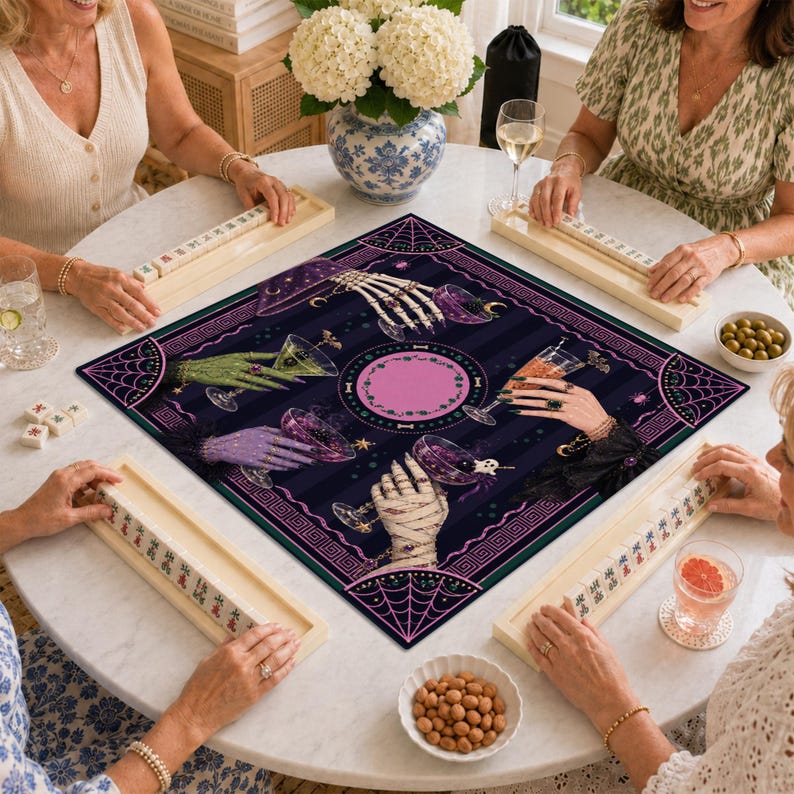 May include: A square game mat featuring a dark purple and black striped background with a central pink circle. The design displays four hands, including skeletal, green, purple, and mummy-wrapped versions, each holding a cocktail glass.