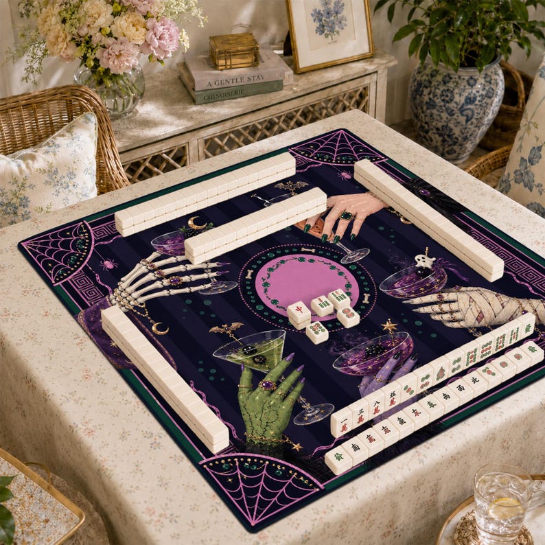May include: A square mahjong mat featuring a dark purple and black striped background with illustrations of skeleton hands, green monster hands, and mummy hands holding cocktail glasses. The mat includes white mahjong tiles and racks.