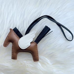 Peut inclure: Un porte-clés en cuir marron en forme de cheval avec une aile en cuir blanc, une crinière et une queue en cuir noir. L'objet comprend une longue lanière en cuir noir pour l'attache, avec des coutures visibles sur les bords du corps du cheval.