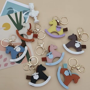 May include: A selection of rocking horse keyrings in various colours. Each keyring features a rocking horse design with a saddle and tassel, attached to a gold-coloured keyring. The keyrings are available in shades of blue, pink, yellow, brown, and black.