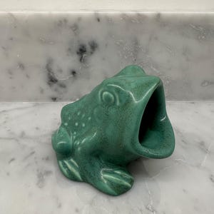 May include: A ceramic frog figurine with a wide, open mouth, finished in a mottled, matte green glaze. The sculpture features textured skin details, including raised bumps on the back and defined limbs, presented in a seated, forward-facing position.