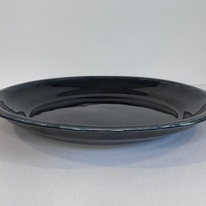 May include: A dark blue ceramic plate with a glossy finish. The plate has a slightly raised rim and a smooth, reflective surface. The plate is round and appears to be empty. The plate is sitting on a white surface.