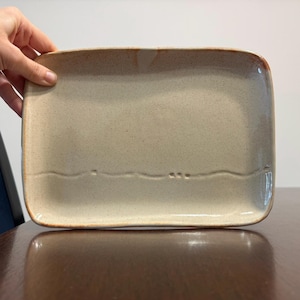 May include: A rectangular ceramic serving platter with a speckled beige finish and a smooth, glossy glaze. The platter features a subtle, horizontal wavy line embossed across the lower section, with a warm, brownish-orange rim accenting the edges.