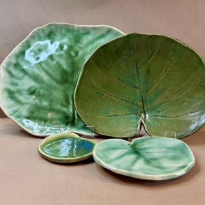 May include: A collection of four ceramic leaf-shaped dishes in varying shades of green. The largest plate is a light green, while the others range from medium to dark green. The plates have detailed vein patterns and are arranged on a neutral surface.