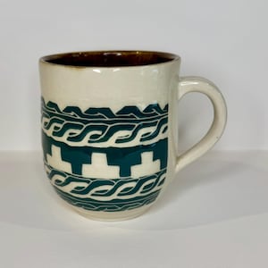 May include: Cream-colored ceramic mug with a dark brown interior and a green geometric pattern. The mug has a curved handle and a cylindrical shape, suitable for hot beverages.