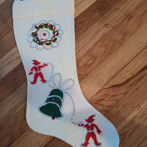 May include: Cream-colored Christmas stocking with festive embellishments. Features a wreath, a green bell, and two red elf figures. The decorations are outlined with silver glitter. A loop is at the top for hanging.