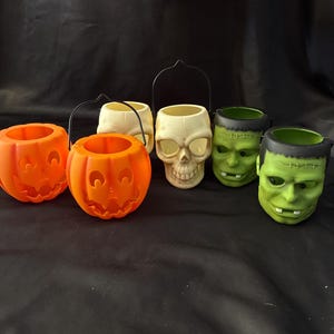 May include: A collection of five plastic Halloween treat buckets featuring two orange jack-o'-lanterns, one off-white skull, and two green monster heads. Each bucket has a hollow interior and a black plastic handle attached to the top rim.