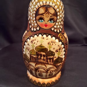 May include: A wooden nesting doll featuring a hand-painted face with blue eyes and a white head covering. The body displays a detailed illustration of a cathedral with golden domes and arches, accented with brown and white decorative patterns.