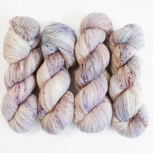 May include: Four skeins of hand-dyed yarn in a neutral color palette. The yarn is a mix of cream, lavender, and burgundy, with subtle speckles throughout. The yarn is arranged on a white background, showcasing its texture and color variations.