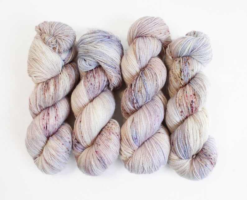 May include: Four skeins of hand-dyed yarn in a neutral color palette. The yarn is a mix of cream, lavender, and burgundy, with subtle speckles throughout. The yarn is arranged on a white background, showcasing its texture and color variations.