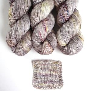 May include: Three skeins of hand-dyed yarn in shades of gray, cream, and hints of yellow and red. A small knitted swatch is also included, showcasing the yarn's color variations. Ideal for knitting and crochet projects.