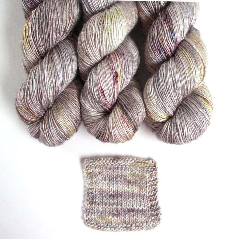 May include: Three skeins of hand-dyed yarn in shades of gray, cream, and hints of yellow and red. A small knitted swatch is also included, showcasing the yarn's color variations. Ideal for knitting and crochet projects.