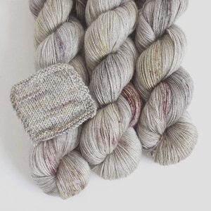 May include: A skein of light gray yarn with subtle color variations, including hints of purple and yellow. A small, knitted swatch is displayed next to the yarn, showcasing the texture and color. Ideal for knitting and crochet projects.