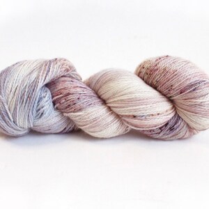 May include: A skein of hand-dyed yarn in a blend of soft colors. The yarn features a mix of white, lavender, and mauve hues, with subtle speckles of darker shades. Ideal for knitting or crochet projects, the yarn is presented against a white background.