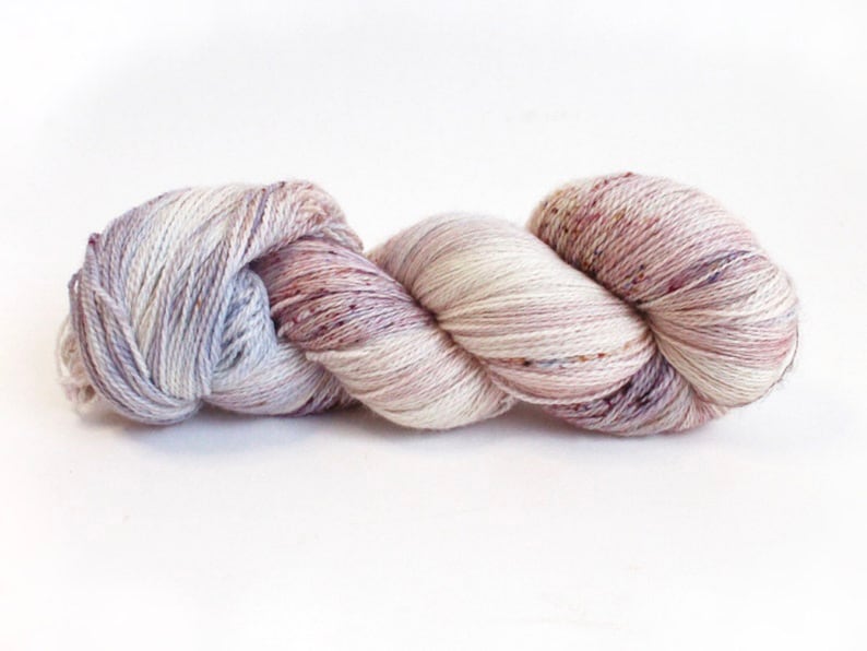 May include: A skein of hand-dyed yarn in a blend of soft colors. The yarn features a mix of white, lavender, and mauve hues, with subtle speckles of darker shades. Ideal for knitting or crochet projects, the yarn is presented against a white background.