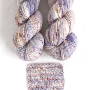 May include: Two skeins of hand-dyed yarn in shades of white, lavender, and brown, with a knitted swatch below. The yarn is twisted into a skein shape, showing the color variations. The swatch displays the yarn's texture and color blend.