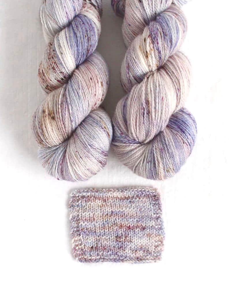 May include: Two skeins of hand-dyed yarn in shades of white, lavender, and brown, with a knitted swatch below. The yarn is twisted into a skein shape, showing the color variations. The swatch displays the yarn's texture and color blend.