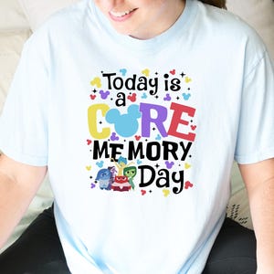 May include: Pale blue t-shirt with the phrase "Today is a CORE MEMORY Day" in vibrant colours and Disney character illustrations. The top has a round neck and short sleeves, suitable for everyday wear.