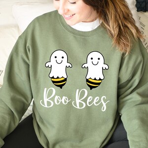 May include: An olive green sweatshirt with a design of two cartoon ghost-bee hybrids. The design features white ghosts with yellow and black striped bee bottoms, and the words "Boo Bees" in a script font.