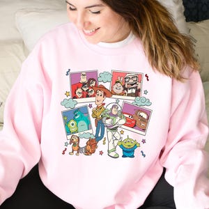 May include: A pale pink sweatshirt with a vibrant graphic of animated characters from Toy Story, Monsters, Inc., and Cars. The design includes images of Woody, Buzz Lightyear, and Sully.
