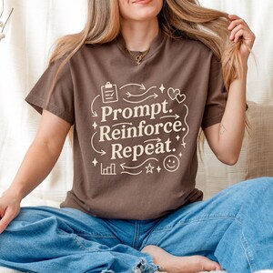 Può includere: Una maglietta marrone con la scritta "Prompt. Reinforce. Repeat." in bianco, con un carattere fantasioso. Il design include frecce, cuori, stelle e una faccina sorridente. La persona indossa jeans blu.
