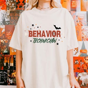 May include: A cream-coloured t-shirt featuring the words "BEHAVIOR TECHNICHAN" in a Halloween design. The design includes a black bat, stars, and orange and green accents. The shirt is a casual, short-sleeved style.