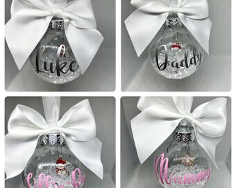 May include: Four clear glass Christmas baubles, each with a white bow and silver top. The baubles are inscribed with names or words: "Luke", "Daddy", "Lillie R", and "Mummy". They are filled with clear beads and have small decorative charms.