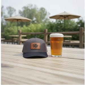 Puede incluir: Gorra de béisbol gris y azul marino con un parche de cuero marrón con un diseño de pájaro, junto a una pinta de cerveza con espuma blanca. Los artículos están sobre una mesa de madera al aire libre.