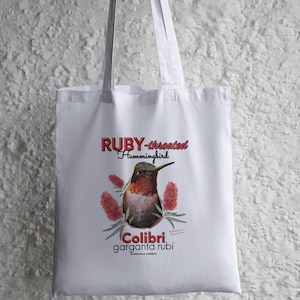 May include: White tote bag featuring a detailed illustration of a Ruby-throated Hummingbird with red and green accents. The text "RUBY-throated Hummingbird" is above the bird, and "Colibri garganta rubí" is below.