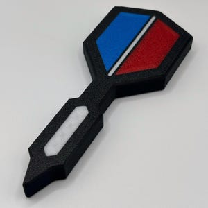 May include: A black plastic dart with a red, white and blue target. The target is shaped like a hexagon.