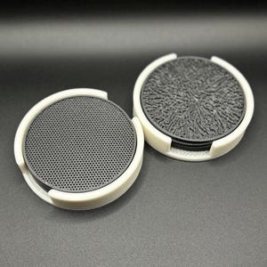 Op de afbeelding: Twee sets zwarte onderzetters met witte randen. De ene set heeft een getextureerd oppervlak en de andere een rasterpatroon.