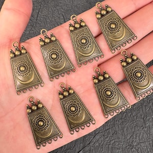 May include: A collection of eight antique bronze-toned metal charms, each featuring a trapezoidal shape with a central circular floral motif above a crescent moon. Each charm includes three small spheres at the top and three loops at the base.