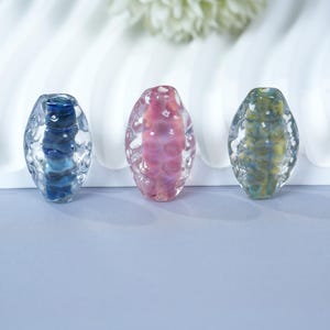 Könnte beinhalten: Drei ovale Glasperlen in Blau, Rosa und Grün. Jede Perle hat eine strukturierte Oberfläche und einen farbigen Kern. Die Perlen werden vor einem weißen Hintergrund präsentiert.