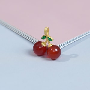 Puede incluir: Un colgante de cereza dorado con dos piedras de cereza redondas y rojas. El colgante tiene un tallo dorado y dos hojas verdes. El colgante está sobre un fondo rosa y azul.