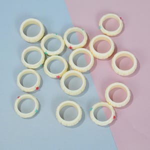May include: A collection of ivory-colored rings with a textured outer edge and small colored accents. The rings are arranged on a two-tone background of pink and blue, creating a visually appealing display. The rings are circular in shape.