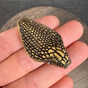 May include: A metallic gold-toned snake head ornament featuring a detailed, raised scale pattern across its surface. The object has an elongated, pointed shape with a smooth, polished finish that highlights the texture of the individual scales.