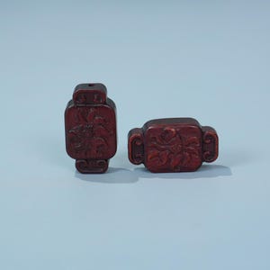 May include: Two dark red, rectangular beads with carved floral designs. Each bead has a hole at the top for stringing. The beads have rounded edges and decorative side elements. The background is a light blue.