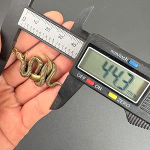 May include: A bronze-colored snake pendant with a crescent moon design is measured with a digital caliper. The digital display reads 44.3 mm. The pendant is held in a hand against a dark background.