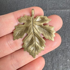 May include: A bronze-toned metal pendant shaped like a grape leaf with a textured surface and defined veins. The leaf features a small loop at the top for attachment, showcasing a metallic finish with dark highlights in the recessed areas.