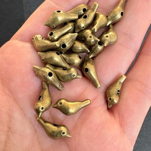 Könnte beinhalten: Eine Sammlung kleiner, antiker Vogelperlen aus Messing. Jede Perle hat ein detailliertes Vogeldesign mit einem Loch zum Auffädeln. Die Perlen haben ein Vintage-Aussehen, ideal für die Schmuckherstellung oder Bastelprojekte.