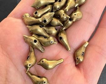 May include: A collection of small, antique brass bird-shaped beads. Each bead features a detailed bird design with a hole for stringing. The beads have a vintage, weathered appearance, perfect for jewelry making or craft projects.