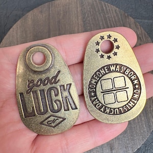 Könnte beinhalten: Zwei antike Metallanhänger in Messingfarbe. Ein Anhänger trägt die Aufschrift "good LUCK" mit einem rautenförmigen Design. Der andere Anhänger hat den Text "SOMEONE WAS BORN ON THIS LUCKY DAY" um ein quadratisches Design mit vier Quadraten und einem Sternenkreis.