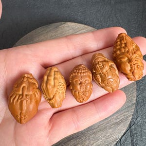 以下が含まれることがあります： さまざまな形とデザインの、小さく精巧に彫刻された木製の置物5つ。彫刻は顔や抽象的な形を描いており、すべて暖かく自然な木の色合いです。置物は手で持たれており、そのサイズと詳細を示しています。