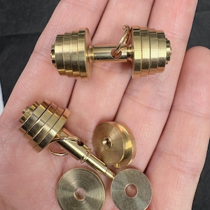 Op de afbeelding: Twee kleine, goudkleurige halterbedels. Eén is volledig gemonteerd, terwijl de andere gedeeltelijk gedemonteerd is, met aparte gewichtsplaten. De halters hebben een klassiek ontwerp, met gestapelde cirkelvormige gewichten aan elk uiteinde en een centrale handgreep.