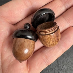 Op de afbeelding: Een kleine houten houder in de vorm van een eikel, bestaande uit een gladde bruine basis en een donkere, gepolijste dop. De houder is in twee delen weergegeven, waarbij de dop is losgemaakt om de holle binnenkant en de schroefdraad te tonen.