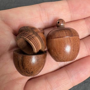 May include: Two small, round, brown wooden containers with a smooth, polished finish. One container is open, revealing a threaded interior rim, while the other is closed and features a small, circular knob at the top centre of its lid.